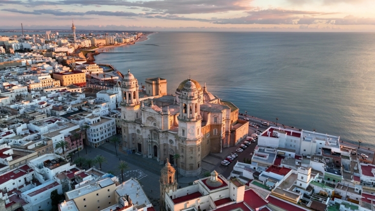 Een luchtfoto van een kerk en omliggende gebouwen in Cádiz bij zonsondergang, met de oceaan op de achtergrond