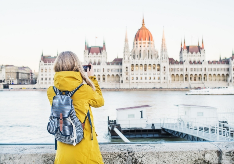 Fille blonde avec manteau jaune et sac à dos rayé prenant une photo de l'édifice parlementaire de Budapest de l'autre côté du Danube